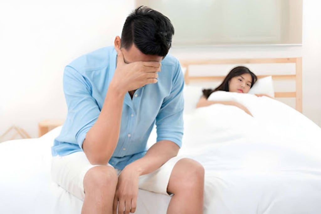 Man sitting on the edge of a bed looking distressed while a woman lies in the background, illustrating erectile dysfunction and relationship stress.