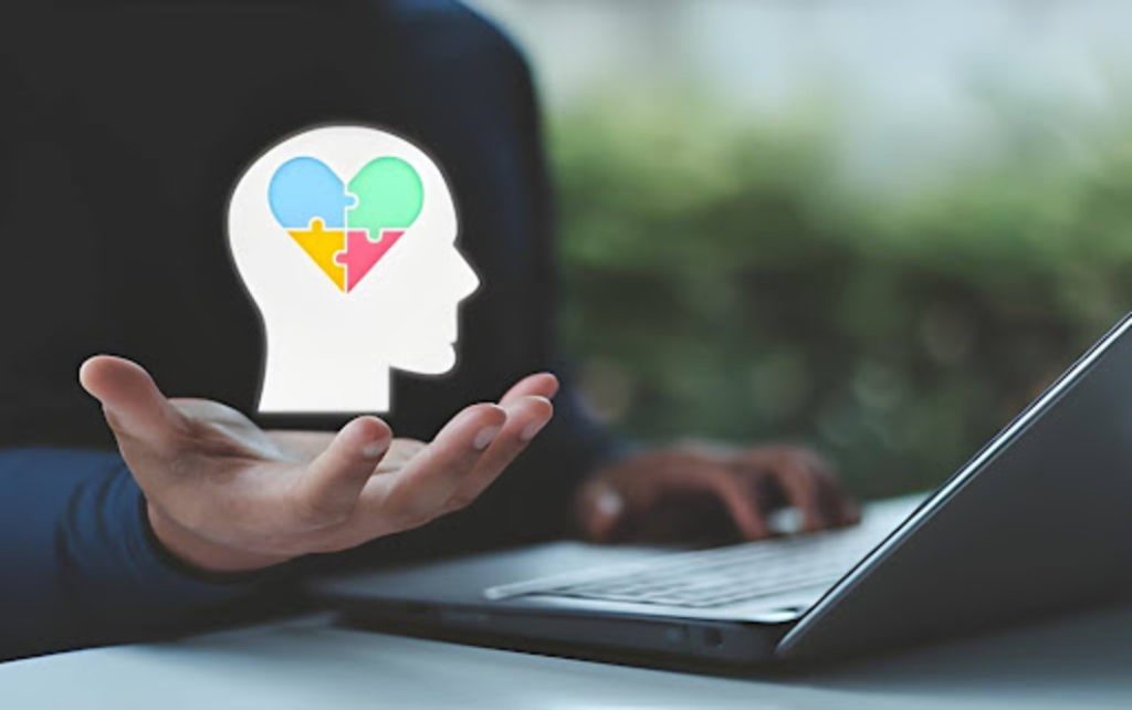Hand holding a mental health icon with colorful puzzle pieces inside a head shape beside an open laptop.
