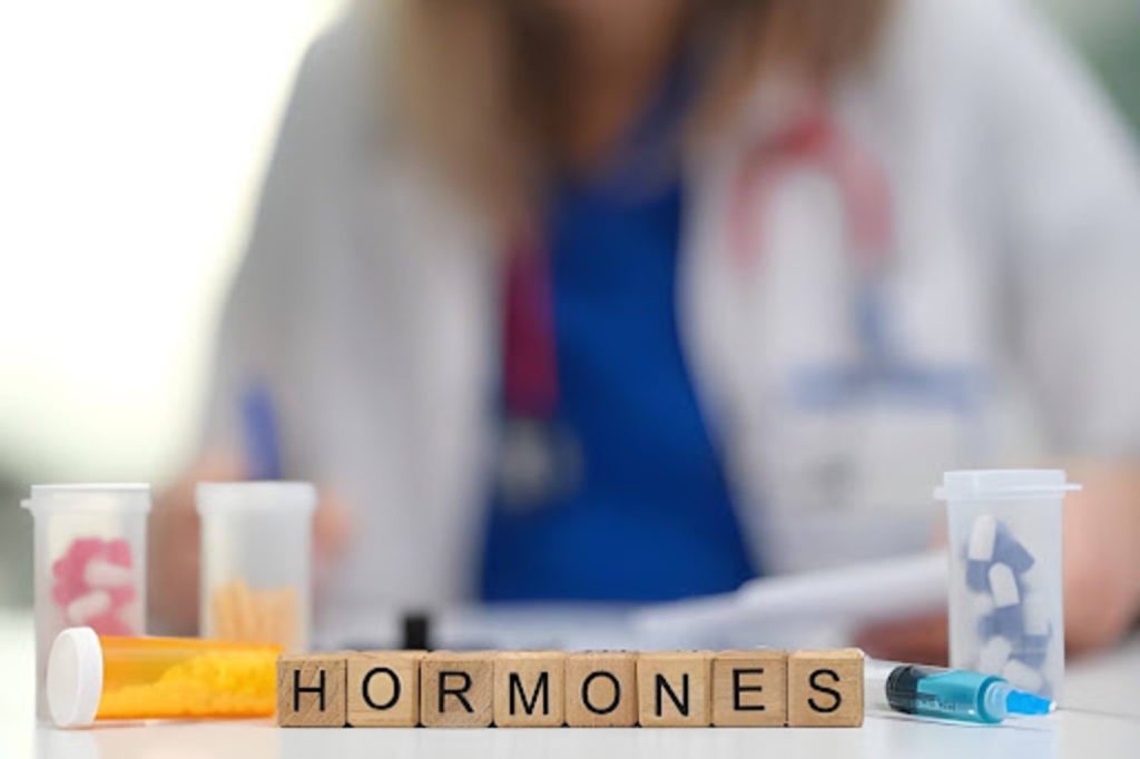 Wooden blocks spelling hormones beside prescription bottles and a syringe with a medical provider in the background.