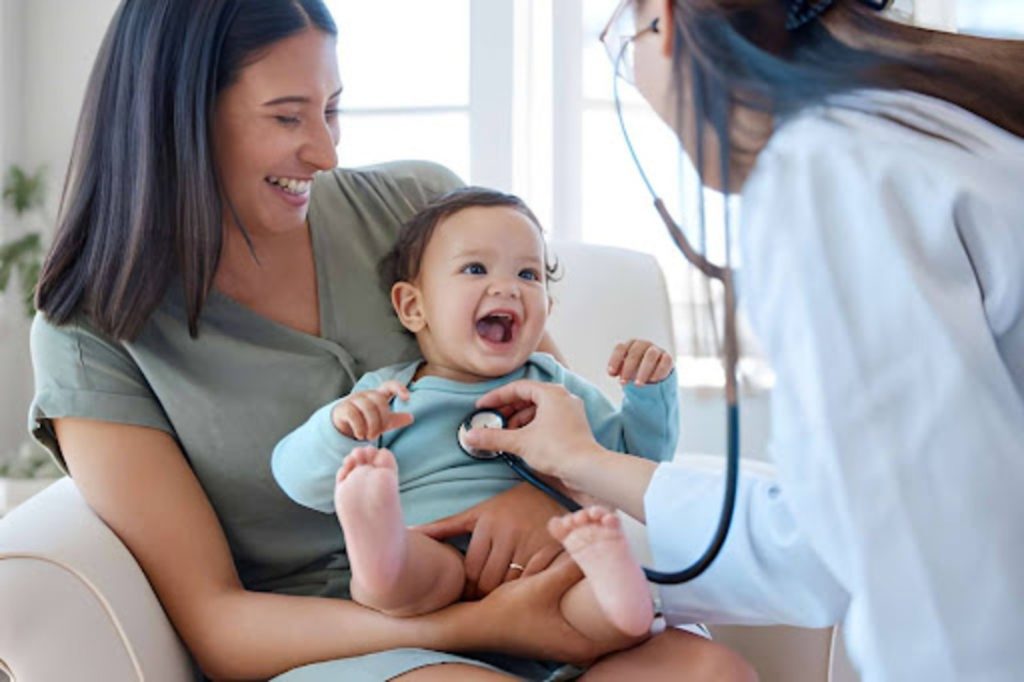 Mother holding a happy baby while a pediatrician listens to the baby’s chest with a stethoscope during a checkup.