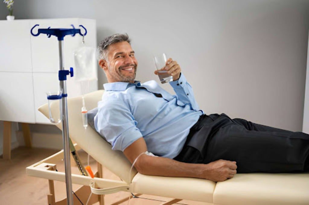 Man reclining on a clinic chair receiving IV therapy, smiling and holding a glass of water while the IV drip infuses fluids into his arm.