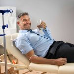 Man reclining on a clinic chair receiving IV therapy, smiling and holding a glass of water while the IV drip infuses fluids into his arm.