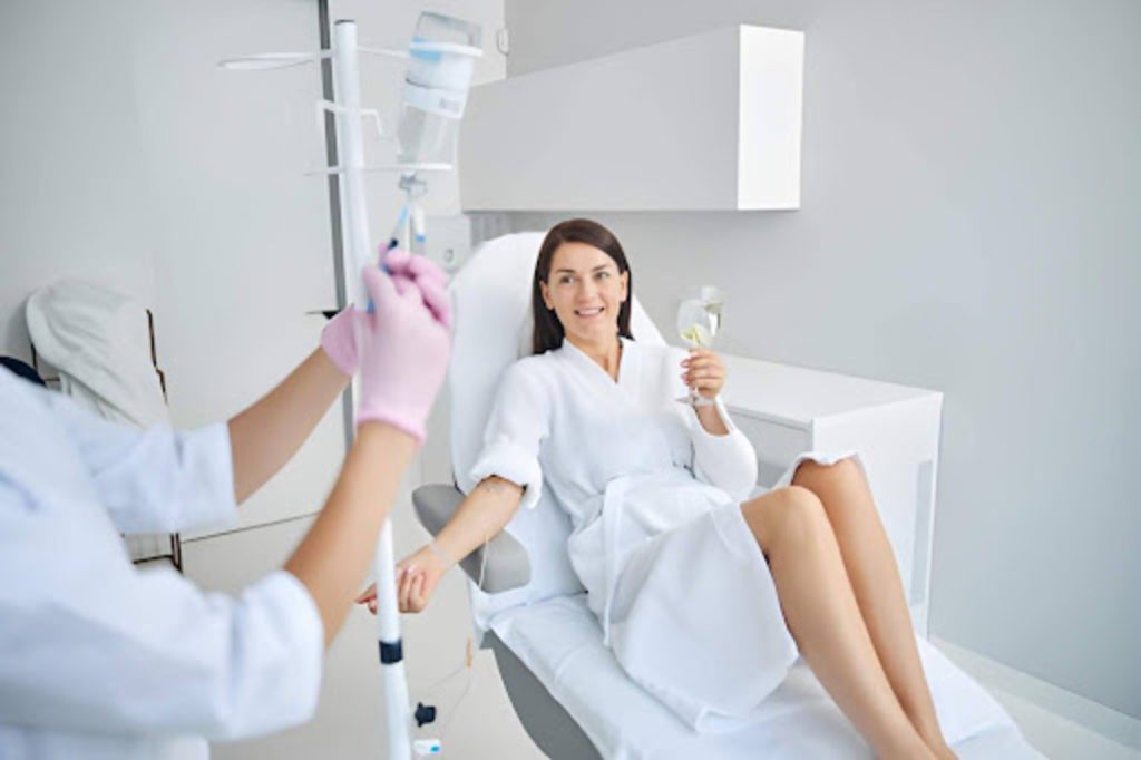 Woman receiving IV therapy while reclining in a clinic chair, holding a glass of water, as a healthcare professional adjusts the IV drip.