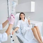 Woman receiving IV therapy while reclining in a clinic chair, holding a glass of water, as a healthcare professional adjusts the IV drip.